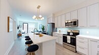 Located at 20 Burling Ln in New Rochelle, NY, this thoughtfully designed single-floor residence seamlessly blends contemporary style with everyday functionality. The open-concept kitchen is a standout centerpiece, featuring crisp white shaker cabinetry, a soft aqua glass tile backsplash, quartz countertops, and stainless steel appliances. A large waterfall-edge island with an integrated sink and bar seating is crowned by a striking gold multi-globe chandelier, making it ideal for both cooking and entertaining. The kitchen flows effortlessly into the living area, which is bathed in natural light from large windows and a glass door leading to an outdoor balcony. The living space offers a comfortable, modern ambiance with light hardwood-style flooring throughout. The bedroom is a serene retreat, highlighted by a bold deep-blue accent wall, large windows that bring in natural light, and clean, neutral walls that complement the warm wood tones of the furnishings. The bathroom features elegant marble-look tile flooring, a glass-enclosed walk-in shower, a wide vanity with white cabinetry, and a bold navy accent wall that adds a sophisticated, cohesive design touch throughout the residence.