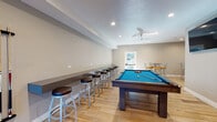 This single-floor recreational space is thoughtfully designed as a full-featured game room and social gathering area. The open layout is unified by light wood-look flooring throughout, recessed lighting, and ceiling fans that maintain a comfortable atmosphere. Neutral gray and taupe wall tones create a clean, modern backdrop for the variety of activities the space accommodates.

The main game area is well-equipped, housing a pool table with a vivid blue felt surface, a regulation shuffleboard table, a foosball table, and a ping-pong table—providing diverse entertainment options. A wall-mounted flat-screen television is positioned prominently for viewing convenience. Cue racks and storage accessories are neatly arranged along the walls near the pool table.

A built-in counter runs along one wall, lined with cushioned bar stools, offering a casual seating area for spectators or socializing. A separate high-top dining nook features round tables paired with colorful padded chairs, adding a vibrant touch to the space.

Multiple exterior doors allow natural light to filter in and provide convenient access to the outdoors. Split-system air conditioning units ensure year-round climate control. The overall design balances functionality with a welcoming, activity-oriented ambiance suited for group recreation.