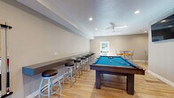 This single-floor recreational space is thoughtfully designed as a full-featured game room and social gathering area. The open layout is unified by light wood-look flooring throughout, recessed lighting, and ceiling fans that maintain a comfortable atmosphere. Neutral gray and taupe wall tones create a clean, modern backdrop for the variety of activities the space accommodates.

The main game area is well-equipped, housing a pool table with a vivid blue felt surface, a regulation shuffleboard table, a foosball table, and a ping-pong table—providing diverse entertainment options. A wall-mounted flat-screen television is positioned prominently for viewing convenience. Cue racks and storage accessories are neatly arranged along the walls near the pool table.

A built-in counter runs along one wall, lined with cushioned bar stools, offering a casual seating area for spectators or socializing. A separate high-top dining nook features round tables paired with colorful padded chairs, adding a vibrant touch to the space.

Multiple exterior doors allow natural light to filter in and provide convenient access to the outdoors. Split-system air conditioning units ensure year-round climate control. The overall design balances functionality with a welcoming, activity-oriented ambiance suited for group recreation.