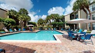 This community amenity space is defined by its resort-style outdoor pool area, offering a welcoming retreat with a distinctly tropical Florida aesthetic. The generously sized swimming pool serves as the centerpiece, with clearly marked shallow-end depth indicators and a surrounding deck finished in decorative paver stone that adds texture and visual warmth to the space. Blue lounge chairs and sun loungers are arranged around the pool deck and along the building's perimeter, providing ample space for relaxation.

The single-story clubhouse building features a stucco exterior with large sliding and swinging glass doors that open directly onto the pool deck, creating a seamless indoor-outdoor connection. White curtains frame the glass entry doors, offering a clean, polished look. The building's entry, marked with an Atlantic Pacific Management mat, is shaded and accessible via a gentle ramp.

Lush tropical landscaping, including mature palm trees, broad-leafed shade trees, and manicured shrubs with red mulch borders, frames the space and creates a private, garden-like atmosphere. Shade umbrellas are positioned around the deck to provide relief from the sun. The surrounding residential buildings, visible beyond the perimeter fencing, complete the enclosed community setting under open Florida skies.