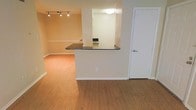 This single-floor residence is designed with an open and functional layout that seamlessly connects the living and kitchen areas. The flooring throughout consists of warm-toned hardwood-style planks, lending a cohesive and inviting feel to the space. The living area is illuminated by a track lighting system mounted on the ceiling, offering adjustable, directed light across the room. Neutral, cream-toned walls complement the overall palette and create a bright, airy ambiance.

A defining architectural feature of this space is the peninsula-style kitchen pass-through, which incorporates a dark granite or stone countertop that extends outward to separate the kitchen from the living area while maintaining visual openness. The kitchen, visible through the pass-through, includes built-in appliances and is lit by a flush-mount ceiling light. White-painted door trim and baseboards frame each wall cleanly, adding a polished finish. A standard interior door with a brushed metal handle is positioned to one side, likely leading to additional rooms or a closet, contributing to the practical flow of the layout.