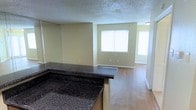 This single-story space features an open-concept design that connects the kitchen and living areas, creating a seamless and functional flow. The kitchen is defined by a prominent dark granite-style bar counter with a wraparound design, offering both workspace and a casual dining or serving area. The kitchen also includes dark cabinetry, adding contrast to the otherwise light and neutral color palette throughout.

The living area extends naturally from the kitchen, finished with warm laminate wood flooring that runs consistently throughout the space. Soft beige walls complement the flooring and contribute to the overall bright and inviting atmosphere. A large window in the living area allows natural light to fill the room, enhancing the sense of openness.

The textured popcorn ceiling is a consistent feature overhead, and white baseboards frame the floors cleanly throughout. A hallway visible from the living area suggests additional rooms beyond, further extending the functional layout of the space. Overall, the unit presents a clean, practical, and well-lit environment suited for comfortable everyday living.