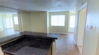 This single-story space features an open-concept design that connects the kitchen and living areas, creating a seamless and functional flow. The kitchen is defined by a prominent dark granite-style bar counter with a wraparound design, offering both workspace and a casual dining or serving area. The kitchen also includes dark cabinetry, adding contrast to the otherwise light and neutral color palette throughout.

The living area extends naturally from the kitchen, finished with warm laminate wood flooring that runs consistently throughout the space. Soft beige walls complement the flooring and contribute to the overall bright and inviting atmosphere. A large window in the living area allows natural light to fill the room, enhancing the sense of openness.

The textured popcorn ceiling is a consistent feature overhead, and white baseboards frame the floors cleanly throughout. A hallway visible from the living area suggests additional rooms beyond, further extending the functional layout of the space. Overall, the unit presents a clean, practical, and well-lit environment suited for comfortable everyday living.