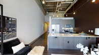 This stylish 1-bedroom, 1-bathroom loft-style residence offers a compelling blend of industrial character and modern design. The open-concept living area immediately impresses with dramatically high ceilings that expose the raw architectural elements above, including visible ductwork, concrete soffits, and pendant track lighting that lend an authentic urban loft aesthetic.

The kitchen serves as a functional centerpiece of the main living space, featuring dark wood cabinetry that extends to the ceiling, a striking marble-topped island with an undermount sink, and a full suite of stainless steel appliances including a French door refrigerator and built-in microwave. The island provides additional seating and workspace, seamlessly connecting the kitchen to the living area.

Rich, dark hardwood flooring runs throughout, grounding the space and complementing the cool tones of the cabinetry and concrete finishes. A hallway leads to the bedroom and bathroom, maintaining a clean, uncluttered flow. The tall, smooth white walls offer a refined contrast to the raw industrial ceiling above. Track lighting throughout provides flexible, layered illumination. The overall ambiance is sophisticated and urban, making this a well-appointed space ideal for modern city living.