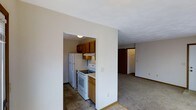 This single-floor space features a straightforward, functional layout with clearly defined areas. The kitchen is a compact, galley-style space enclosed by walls on either side, equipped with a white electric range, a full-size white refrigerator, and an under-cabinet range hood. Medium-toned honey oak cabinetry lines the upper portion of the kitchen walls, providing ample storage. The flooring in the kitchen consists of tile in earthy, muted tones, offering a practical and easy-to-maintain surface.

Adjacent to the kitchen, the living area opens up with wall-to-wall carpeting in a neutral beige tone, contributing to a soft, understated atmosphere. The white walls throughout the space create a clean, bright backdrop, while flush-mount ceiling lighting provides adequate illumination. A doorway visible from the living area leads further into the unit, suggesting additional rooms beyond. Baseboards in white trim line the perimeter of the rooms, adding a subtle finishing detail. Overall, the space presents a simple, utilitarian design with a neutral palette suited for practical, everyday living.