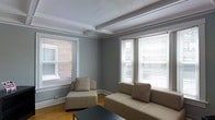 This single-floor space combines classic architectural character with a clean, contemporary feel. The living area immediately draws attention with its distinctive coffered ceiling, detailed with white-painted beams that add a sense of structure and elegance overhead. A textured ceiling finish further contributes to the room's traditional charm.

Two generously sized windows, dressed with horizontal blinds, allow abundant natural light to pour into the room from multiple angles, creating a bright and welcoming atmosphere. Views through the windows reveal a brick exterior neighboring the space, lending an urban character to the setting. The crisp white window trim and baseboards provide a sharp contrast against the cool gray walls, reinforcing a clean and polished aesthetic.

The hardwood flooring anchors the space with warmth and continuity, complementing the neutral color palette throughout. The living area is furnished with a modern sectional sofa and a matching accent chair, both in a warm tan upholstery accented with patterned throw pillows, creating a comfortable and cohesive seating arrangement. A minimalist dark coffee table completes the setup.

Overall, the space offers a harmonious blend of timeless architectural details and modern furnishings, making it both functional and inviting.