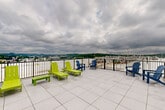Located at 19 Hemans St in Worcester, MA, this rooftop amenity space combines a comfortable indoor lounge with a spacious outdoor terrace, offering residents a well-rounded retreat above the city. The interior lounge features neutral-toned walls, striped carpeting in shades of blue and gray, and modern seating, creating a relaxed and welcoming atmosphere. A stainless steel elevator provides convenient access to the floor, while large glass doors and windows flood the space with natural light and offer a seamless visual connection to the outdoors. A foosball table adds a recreational touch, making the lounge an inviting gathering spot. Transitioning outside, the expansive rooftop terrace is finished with large-format tile flooring and enclosed by a sleek metal railing. Vibrant lime green chaise lounges and blue Adirondack chairs are arranged to take full advantage of the panoramic views of the surrounding hills and Worcester skyline. The open-air terrace is an ideal setting for relaxing or socializing, with ample space for multiple seating arrangements and an outdoor shower station for added convenience. Together, the indoor and outdoor areas create a cohesive and functional rooftop amenity experience.