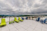 Located at 19 Hemans St in Worcester, MA, this rooftop amenity space combines a comfortable indoor lounge with a spacious outdoor terrace, offering residents a well-rounded retreat above the city. The interior lounge features neutral-toned walls, striped carpeting in shades of blue and gray, and modern seating, creating a relaxed and welcoming atmosphere. A stainless steel elevator provides convenient access to the floor, while large glass doors and windows flood the space with natural light and offer a seamless visual connection to the outdoors. A foosball table adds a recreational touch, making the lounge an inviting gathering spot. Transitioning outside, the expansive rooftop terrace is finished with large-format tile flooring and enclosed by a sleek metal railing. Vibrant lime green chaise lounges and blue Adirondack chairs are arranged to take full advantage of the panoramic views of the surrounding hills and Worcester skyline. The open-air terrace is an ideal setting for relaxing or socializing, with ample space for multiple seating arrangements and an outdoor shower station for added convenience. Together, the indoor and outdoor areas create a cohesive and functional rooftop amenity experience.