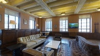 This single-floor space showcases a striking blend of historic architectural craftsmanship and contemporary furnishings. The most notable feature is the elaborately detailed coffered ceiling, adorned with decorative moldings and floral motifs, painted in muted gold and cream tones that draw the eye upward and contribute to an air of grandeur. Pendant light fixtures hang from the ceiling's recessed panels, casting a warm ambient glow throughout the room.

The walls are finished in a rich, warm terracotta tone, complemented by dark wood wainscoting that adds depth and traditional elegance. Multiple windows along the perimeter flood the interior with natural light while offering views of an urban skyline, reinforcing the space's city setting.

The lounge area is anchored by a large area rug featuring intricate patterns, upon which a collection of tufted and structured leather sofas and armchairs are arranged around a sleek, low-profile coffee table. A flat-screen television is mounted on one wall, providing a modern counterpoint to the space's classical detailing. Dark hardwood flooring ties the room together, enhancing its warm, sophisticated ambiance. Overall, the space feels refined, comfortable, and rich in architectural character.