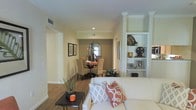 This single-floor residence offers a cohesive and inviting layout designed for comfortable living. The living area serves as the heart of the home, featuring a plush white sofa accented with coral and patterned throw pillows, warm hardwood flooring, and a sleek dark coffee table. A striking built-in shelving unit with white trim adds both function and character, displaying decorative accents that lend a personalized touch to the space. Crown molding runs throughout, adding a refined architectural detail that elevates the overall aesthetic. Recessed lighting illuminates the interiors, complementing the natural warmth of the neutral color palette that flows seamlessly from room to room. The dining area, visible from the living space, features a round glass-top table surrounded by woven rattan-style chairs, creating an elegant yet relaxed atmosphere. A large arched mirror on the accent wall adds depth and visual interest to the dining space. Hallways connect the areas fluidly, reinforcing the open and airy feel of the floor plan. Decorative greenery is thoughtfully placed throughout, bringing a touch of nature indoors. Overall, this home balances classic architectural details with warm, transitional décor for a welcoming and polished living environment.