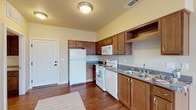 This single-floor unit offers a practical and comfortable layout designed for everyday living. The kitchen is a standout feature, equipped with warm medium-toned wood cabinetry extending to the upper walls, providing generous storage throughout. Dark gray countertops complement the cabinetry, while white appliances—including a refrigerator, electric range, over-the-range microwave, and dishwasher—complete the space. Rich hardwood-style flooring anchors the kitchen area, adding warmth and visual contrast to the light-toned walls.

The kitchen opens directly into the living area, which transitions to plush neutral carpeting, creating a clear and comfortable delineation between spaces. A large window fitted with vertical blinds allows natural light to fill the living area, contributing to a bright and airy atmosphere. A sliding glass door provides additional light and outdoor access.

The bedroom is a well-proportioned room featuring soft carpeted flooring, neutral walls, and a window with vertical blinds that brings in natural light. The space offers a calm and restful environment.

The bathroom includes a tub-shower combination, wood-toned cabinetry with a light countertop, and hardwood-style flooring that ties back to the rest of the unit's design. Overall, the space maintains a cohesive, functional aesthetic throughout.