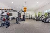 Located at 2800 Milan Ct in Birmingham, Alabama, this fitness center offers a generously sized, single-floor exercise space designed to support a comprehensive range of workout routines. The room features soaring ceilings and an abundance of natural light streaming through large windows that line the walls, creating an open, energizing atmosphere with outdoor views.

The cardio zone is well-appointed with multiple treadmills, stair climbers, ellipticals, and stationary bikes, all positioned to take advantage of the natural light and outdoor scenery. The strength training area includes a robust selection of Matrix selectorized machines for targeted muscle training, along with a multi-functional rack system equipped with battle ropes, resistance attachments, and storage for medicine balls and kettlebells. A dedicated free weight section features a full rack of dumbbells for varied training needs.

A convenient amenity station with dark cabinetry, overhead storage, a beverage refrigerator, and a water dispenser provides easy access to hydration. Wall-mounted flat-screen TVs are positioned throughout the space for entertainment during workouts. Pendant lighting fixtures and recessed lighting complement the natural light, while ceiling fans help maintain a comfortable environment. The durable, speckled flooring throughout is both practical and polished, reinforcing the facility's modern, well-maintained aesthetic.