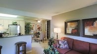 This single-floor residence offers a thoughtfully designed open-concept layout that encourages a natural flow between the living, dining, and kitchen areas. The living room serves as the heart of the space, furnished with a plush sofa accented by decorative throw pillows and complemented by a classic table lamp that casts a warm ambient glow. A large framed artwork adds a bold focal point to the neutral-toned walls, enhancing the room's refined aesthetic. Soft wall-to-wall carpeting throughout the main living and dining areas contributes to a cozy, comfortable atmosphere. The dining area, positioned adjacent to the living room, features a dark wood table surrounded by wicker-style chairs beneath a ceiling fan with integrated lighting, creating an intimate setting for meals. A built-in shelving unit provides both functional storage and a visual divider between spaces. The kitchen is partially open to the main living area via a breakfast bar with bar stool seating, making it ideal for casual dining or entertaining. Dark countertops contrast with light cabinetry for a balanced, timeless look. Potted greenery throughout the space adds life and warmth, enhancing the overall inviting ambiance of this well-composed residence.