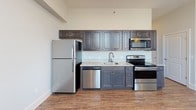 Located at 120 W Saint John St in Spartanburg, South Carolina, this single-floor unit offers a well-appointed, open-concept layout with a clean, modern aesthetic throughout. The kitchen serves as a functional centerpiece, featuring dark espresso cabinetry extending to the upper walls, light granite countertops, and a full suite of stainless steel appliances including a refrigerator, electric range, built-in microwave, and dishwasher. A single-basin sink with a contemporary faucet anchors the counter space, providing a practical and polished workspace.

Hardwood-style flooring runs consistently throughout the unit, lending warmth and visual continuity from the kitchen into the open living area. The living space is generously proportioned, with neutral gray-toned walls and large windows fitted with horizontal blinds that allow abundant natural light and offer elevated views of the surrounding Spartanburg cityscape. Wall-mounted sconces add accent lighting, while a wall-mounted mini-split HVAC unit ensures year-round climate control.

Interior doors feature classic raised-panel designs, maintaining a refined yet understated character throughout. The bathroom includes a tub-shower combination with a white surround, dark vanity cabinetry with granite countertops, and matching hardwood-style flooring, completing the cohesive design palette of the unit.