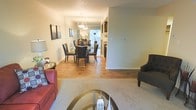 This single-floor residence offers a thoughtfully designed open-concept layout that seamlessly connects the living and dining areas, creating a cohesive and functional living environment. The living room features plush carpeting, providing a comfortable and inviting atmosphere, and is furnished with classic seating arrangements centered around a glass-topped coffee table. A transition to warm hardwood-style flooring marks the flow into the dining area, where a round glass dining table surrounded by dark leather chairs serves as an elegant focal point. A pendant light fixture with a geometric design hangs above the dining table, adding a stylish touch to the space. Natural light filters in through a window at the far end of the dining area, brightening the room and enhancing the overall sense of openness. The neutral-toned walls throughout provide a clean, versatile backdrop that complements the warm flooring tones. Beyond the dining area, a glimpse of the kitchen reveals additional cabinetry and countertop space. The layout and design of this residence offer a harmonious balance of comfort and style, making it well-suited for both everyday living and entertaining.