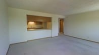 This single-floor space showcases an open and functional layout designed to maximize usability. The living area is generously sized, finished with wall-to-wall neutral carpeting that adds warmth underfoot. Crisp white walls throughout create a bright, clean atmosphere.

A defining architectural feature is the pass-through kitchen window, which opens directly into the living area, promoting a connected and sociable feel between the two spaces. The kitchen is equipped with wooden cabinetry in a warm medium-brown tone, offering ample storage. Under-cabinet lighting illuminates the countertop workspace, and a sink with a standard faucet is positioned beneath the pass-through opening. A cooktop is also visible along the counter, rounding out the kitchen's core functionality.

A hallway visible from the living area leads further into the unit, suggesting additional rooms beyond. The space maintains a neutral, move-in-ready aesthetic with consistent trim and baseboards throughout. The overall ambiance is practical and understated, well-suited for everyday living with a straightforward, efficient floor plan.