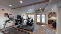 Located at 4569 Driver Lane in Coeur d'Alene, Idaho, this single-floor community facility is designed to support both active wellness and everyday convenience. The space is organized into distinct functional zones, each thoughtfully appointed for its intended purpose.

The fitness area serves as the centerpiece of the facility, divided into two connected sections. The main cardio room features multiple True-brand treadmills and elliptical machines arranged on patterned carpet flooring, with recessed lighting overhead and a wall-mounted TV station. French doors with glass panels provide natural light and direct access to the exterior. A dedicated strength training annex houses a multi-station cable machine and weight equipment, complemented by a full-length wall mirror and carpet flooring, offering a focused environment for resistance training.

A transitional hallway area with carpeted flooring connects the fitness zones to the rest of the facility, featuring wood-tone doors and interior windows that allow visibility between spaces.

The kitchen is well-equipped with rich cherry wood cabinetry, light stone countertops, a black appliance suite including a range and refrigerator, and a central island with a double stainless steel sink illuminated by pendant lighting. Warm hardwood-style flooring flows throughout the kitchen and into the adjacent dining nook, which features ample natural light from surrounding windows—an inviting area for casual seating and meals.