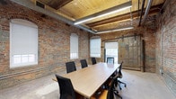 This conference room offers a compelling fusion of historic industrial character and contemporary workspace design. The exposed brick walls, rich with texture and warmth, serve as a defining architectural feature, lending the room a sense of authenticity and heritage. Overhead, natural wood-planked ceilings with visible beams add warmth and contrast, complemented by sleek linear pendant lighting that ensures bright, even illumination throughout. A large rectangular wood-top conference table anchors the space, surrounded by ergonomic task chairs that comfortably accommodate a full team. Natural light filters in through arched windows fitted with roller shades, allowing for flexible light control during presentations or video calls. A prominent industrial-style sliding barn door provides both a functional and aesthetic focal point, hinting at the building's storied past while offering a view to an adjacent area. Exposed ductwork runs along the ceiling, reinforcing the space's industrial aesthetic without detracting from its professional atmosphere. The polished concrete floor ties all elements together, creating a cohesive, modern-industrial ambiance that is both inspiring and highly functional for collaborative work and meetings.
