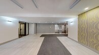 This building lobby is a well-appointed, single-level space offering a comfortable and functional entry experience. Visitors are welcomed through automatic sliding glass doors framed in sleek black metal, which open into a bright, open foyer. The entrance zone is defined by a standout ceiling feature composed of geometric hexagonal panels with recessed lighting, lending a contemporary architectural character to the space.

The flooring throughout is finished in large-format light grey tiles, complemented by grey carpet runner mats that guide traffic from the entrance toward the interior. The lobby transitions seamlessly into an elevator hall, where two stainless steel elevator doors are set against a textured grey brick accent wall, adding warmth and visual depth. Wall-mounted sconces provide supplementary lighting along this corridor.

To one side of the entry, a full wall of individual mailboxes serves residents, positioned adjacent to a decorative feature wall with a gold patterned wallcovering that adds a refined touch. A secondary door provides access to an inner hallway leading further into the building.

The overall ambiance is clean, modern, and welcoming, with neutral tones, recessed lighting, and thoughtful design details creating a polished and practical communal entrance area.