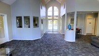 This single-floor space presents an impressive lobby or common area defined by its striking architectural character. The centerpiece is a dramatic vaulted ceiling with exposed beams that rises to a commanding height, amplifying the sense of openness and grandeur. A large arched window positioned at the far end of the entryway floods the interior with abundant natural light, creating a bright and airy ambiance throughout. Double glass doors beneath the arch provide a welcoming entry point and reinforce the connection to the outdoors. The flooring features a multi-toned patterned commercial carpet in neutral and blue-grey hues, adding texture and visual depth to the expansive floor plan. Rounded architectural columns frame the reception or lounge area to the right, where a decorative blue vase and built-in counter surface add a refined, curated touch. To the left, interior windows offer a glimpse into adjacent rooms or offices, suggesting a multi-functional layout suited for administration or resident services. The walls are finished in a clean, crisp white, enhancing the brightness and providing a neutral backdrop that complements the space's functional yet inviting design. Overall, the space conveys a professional and welcoming environment well-suited for a community clubhouse or similar facility.
