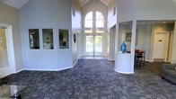 This single-floor space presents an impressive lobby or common area defined by its striking architectural character. The centerpiece is a dramatic vaulted ceiling with exposed beams that rises to a commanding height, amplifying the sense of openness and grandeur. A large arched window positioned at the far end of the entryway floods the interior with abundant natural light, creating a bright and airy ambiance throughout. Double glass doors beneath the arch provide a welcoming entry point and reinforce the connection to the outdoors. The flooring features a multi-toned patterned commercial carpet in neutral and blue-grey hues, adding texture and visual depth to the expansive floor plan. Rounded architectural columns frame the reception or lounge area to the right, where a decorative blue vase and built-in counter surface add a refined, curated touch. To the left, interior windows offer a glimpse into adjacent rooms or offices, suggesting a multi-functional layout suited for administration or resident services. The walls are finished in a clean, crisp white, enhancing the brightness and providing a neutral backdrop that complements the space's functional yet inviting design. Overall, the space conveys a professional and welcoming environment well-suited for a community clubhouse or similar facility.