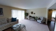 This single-floor space features a well-appointed living room that serves as its central gathering area. The room is finished with wall-to-wall carpet in a neutral beige tone, contributing to a warm and inviting atmosphere throughout. Large sliding glass doors span much of one wall, dressed with vertical blinds that allow for adjustable natural light, brightening the interior and lending an airy, open quality to the space.

The living area is generously proportioned, offering ample room for multiple seating arrangements. A plush sofa with accent pillows anchors one side of the room, paired with a glass-topped coffee table featuring ornate metal legs. Across the room, a cushioned armchair provides additional seating. A classic dark wood writing desk and chair occupy a corner, adding a functional element alongside the decorative framed artwork displayed on the walls.

Potted plants placed near the sliding doors and in the corners introduce natural greenery that softens the space. The walls are finished in a neutral tan, complementing the light-toned ceiling and carpet. Overall, the space presents a clean, comfortable, and practical living environment with a traditional aesthetic sensibility.