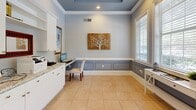 This thoughtfully designed office space offers a refined and functional environment suited for professional use. The room is finished with a distinctive tray ceiling painted in a soft blue-gray hue, accented by recessed lighting and crisp crown molding that adds a polished architectural touch. Walls are adorned in a cool light gray tone, complemented by classic wainscoting with raised panel detailing that lines the lower half of the room, creating a sophisticated visual contrast.

Extensive built-in white cabinetry lines the walls, featuring elegant granite countertops with a soft white-and-gray pattern. Upper cabinets with open shelving provide ample storage, while lower cabinets with brushed nickel hardware offer additional organization. Multiple workstation areas are integrated into the cabinetry, providing dedicated space for computers and office equipment.

Large windows dressed with white horizontal blinds flood the room with natural light and offer views of the surrounding greenery outdoors. Light beige ceramic tile flooring ties the room together with a clean, neutral foundation. A glass-paned door connects the office to an adjacent space, enhancing accessibility. Decorative artwork and a small console table near the windows complete the inviting, professional ambiance of this well-equipped office.