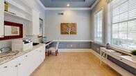 This thoughtfully designed office space offers a refined and functional environment suited for professional use. The room is finished with a distinctive tray ceiling painted in a soft blue-gray hue, accented by recessed lighting and crisp crown molding that adds a polished architectural touch. Walls are adorned in a cool light gray tone, complemented by classic wainscoting with raised panel detailing that lines the lower half of the room, creating a sophisticated visual contrast.

Extensive built-in white cabinetry lines the walls, featuring elegant granite countertops with a soft white-and-gray pattern. Upper cabinets with open shelving provide ample storage, while lower cabinets with brushed nickel hardware offer additional organization. Multiple workstation areas are integrated into the cabinetry, providing dedicated space for computers and office equipment.

Large windows dressed with white horizontal blinds flood the room with natural light and offer views of the surrounding greenery outdoors. Light beige ceramic tile flooring ties the room together with a clean, neutral foundation. A glass-paned door connects the office to an adjacent space, enhancing accessibility. Decorative artwork and a small console table near the windows complete the inviting, professional ambiance of this well-equipped office.