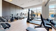 Located at 2475 Old Bronte Rd in Oakville, Ontario, this contemporary fitness facility is thoughtfully designed to support a full range of exercise disciplines. The space is divided into two distinct zones connected by glass-paneled sliding doors, creating an open yet organized layout with light hardwood flooring throughout.

The main gym area is bright and airy, benefiting from expansive floor-to-ceiling windows that flood the space with natural light and offer views of the surrounding neighbourhood. This zone is well-appointed with a comprehensive selection of cardio machines — including treadmills, ellipticals, and stationary bikes — as well as a multi-station cable system, a weight bench, and a full dumbbell rack accommodating a wide range of free weights. Warm wood-toned cabinetry accents the walls, complemented by recessed lighting for a clean, modern aesthetic.

The second area serves as a dedicated functional training studio. It features floor-to-ceiling mirrors that enhance the sense of space, hanging heavy punching bags for boxing and combat training, TRX suspension straps mounted to the wall, and stacked balance and agility equipment. This room offers ample open floor space for stretching, mat work, and dynamic movement exercises.

Together, the two zones form a cohesive, professionally outfitted fitness centre suited for strength training, cardio, and functional fitness alike.