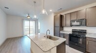 Located at 6405 N Interstate 35 in Denton, TX, this 2-bedroom, 2-bathroom unit offers a thoughtfully designed single-floor layout. The open-concept kitchen and living area serves as the heart of the home, anchored by a large granite-topped island with an undermount sink and stylish pendant lighting. Rich dark brown cabinetry lines the kitchen walls, complemented by matching granite countertops and black appliances, including a refrigerator, range, microwave, and dishwasher. Light wood-style flooring flows throughout the main living areas, while sliding glass doors open to a private balcony, filling the space with natural light and offering outdoor views. Both bedrooms are carpeted and well-lit with windows overlooking the surrounding landscape, each served by a ceiling fan. The primary bedroom connects to a full bathroom featuring granite vanity counters, dark cabinetry, a tiled tub-shower combination, and access to a walk-in closet outfitted with wire shelving systems. The second bathroom is equally well-appointed. Additional conveniences include a dedicated laundry area, utility room, and multiple closets throughout, ensuring ample storage across the entire floor plan.