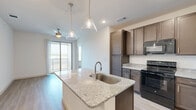 Located at 6405 N Interstate 35 in Denton, TX, this 2-bedroom, 2-bathroom unit offers a thoughtfully designed single-floor layout. The open-concept kitchen and living area serves as the heart of the home, anchored by a large granite-topped island with an undermount sink and stylish pendant lighting. Rich dark brown cabinetry lines the kitchen walls, complemented by matching granite countertops and black appliances, including a refrigerator, range, microwave, and dishwasher. Light wood-style flooring flows throughout the main living areas, while sliding glass doors open to a private balcony, filling the space with natural light and offering outdoor views. Both bedrooms are carpeted and well-lit with windows overlooking the surrounding landscape, each served by a ceiling fan. The primary bedroom connects to a full bathroom featuring granite vanity counters, dark cabinetry, a tiled tub-shower combination, and access to a walk-in closet outfitted with wire shelving systems. The second bathroom is equally well-appointed. Additional conveniences include a dedicated laundry area, utility room, and multiple closets throughout, ensuring ample storage across the entire floor plan.