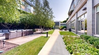 Located in Chicago's vibrant 60647 neighborhood, this thoughtfully designed outdoor courtyard offers a serene urban retreat enclosed by a modern multi-story building with a contemporary façade of brick, glass, and paneling. A wide concrete walkway runs the length of the space, flanked by manicured lawns, mature birch trees, and well-tended garden beds filled with lush shrubs and perennials that provide natural shade and greenery throughout.

The courtyard is divided into several distinct functional zones. A lounge area features comfortable wicker and upholstered outdoor sofas, ideal for casual gathering and relaxation. Nearby, a dedicated outdoor dining area is outfitted with a sleek table and chairs, positioned alongside a built-in grilling station with stone surround — perfect for al fresco dining and entertaining. Additional seating arrangements with lounge chairs and side tables offer versatile spots to unwind in the open air.

Large floor-to-ceiling windows and glass double doors along the building's ground-level facade allow natural light to flow both into the interior and back out into the courtyard, maintaining a seamless indoor-outdoor connection. Bollard lighting is integrated throughout the walkways, ensuring the space is both functional and inviting after dark. The overall ambiance is fresh, open, and well-appointed, offering a peaceful escape within the heart of the city.
