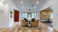 This single-floor community space is thoughtfully designed to provide a comfortable and functional environment for gathering and relaxation. The entry lounge sets a welcoming tone with soaring ceilings, light hardwood-style flooring, and large floor-to-ceiling windows and glass doors that frame lush, tree-lined outdoor views. A seating arrangement of upholstered armchairs centered around a glass-top table, paired with a patterned area rug, creates an inviting focal point. An elegant arched mirror and a tufted bench add refined decorative touches near the entryway.

Adjacent to the lounge, a spacious multi-purpose gathering room features plush carpeting and recessed lighting. It is outfitted with industrial-style bar tables and stools, a wall-mounted flat-screen television, and a cozy seating nook with patterned armchairs—making it ideal for both casual socializing and organized activities. A built-in red wood cabinetry unit provides additional functionality in this room.

The kitchen is a standout feature, showcasing rich cherry wood cabinetry with crown molding detail, white countertops, and light hardwood-style flooring. It includes a full-size refrigerator, microwave, and sink, and benefits from abundant natural light through large windows overlooking the surrounding greenery. The space seamlessly blends practicality with warm, inviting aesthetics throughout.