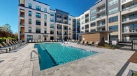 Located at 901 St Albans Dr in Raleigh, NC, this modern multi-story community showcases a contemporary architectural design blending light brick exteriors with bold dark-framed windows, private balconies, and clean geometric lines. The outdoor amenity areas are a standout feature, anchored by a generously sized resort-style swimming pool with a wide stone-paved deck lined with lounge chairs, offering an inviting atmosphere under open skies with wooded views beyond the property.

The interior courtyard provides a well-landscaped retreat with manicured turf lawns, paved walkways, raised planters, and outdoor seating areas equipped with grills and dining furniture — ideal for social gatherings. String lights overhead add a warm ambiance to the courtyard evenings.

A covered ground-floor lounge area features exposed concrete ceilings, whitewashed brick walls, and tile flooring, creating an industrial-chic aesthetic. This space is equipped with comfortable wicker and upholstered seating arranged around a flat-screen television, as well as a wet bar with navy cabinetry — a versatile space for residents to gather and entertain.

Paved corridors connect the various outdoor zones, weaving through thoughtfully planted garden beds and past warm wood-clad accent structures. The two-floor layout ensures convenient access throughout the community, and the abundance of natural light throughout the shared spaces enhances the overall open and welcoming character of this Raleigh property.