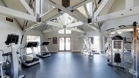 This fitness center is a thoughtfully designed workout facility that combines architectural character with practical functionality. The most striking feature of the space is its soaring vaulted ceiling, supported by an impressive framework of exposed white-painted timber trusses and steel connectors that lend the room an open, airy feel and a distinct rustic-industrial aesthetic.

Natural light floods the interior through multiple windows and a set of elegant French doors, which also provide direct access to the outdoors. The generous ceiling height enhances the sense of spaciousness, making the environment feel expansive and welcoming rather than confined.

The workout floor is finished with durable dark rubber flooring, ideal for high-impact exercise and equipment use. The facility is equipped with cardio machines, including treadmills, as well as strength and resistance training equipment to support a full range of workout routines. Wall-mounted mirrors along one side of the room help users monitor their form while also visually expanding the space.

The neutral gray walls keep the environment calm and focused, complementing the clean white structural elements above. Overall, this fitness center offers a bright, spacious, and well-appointed setting that balances architectural interest with a fully functional exercise environment.