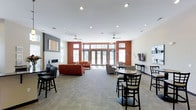 This single-floor communal lounge is designed to accommodate a variety of activities, from casual gatherings to quiet relaxation. The space is defined by striking terracotta-red accent walls that contrast with the neutral white ceiling and light-toned carpeted flooring, creating a warm yet contemporary atmosphere. Recessed lighting throughout the ceiling is complemented by dual ceiling fans, ensuring consistent illumination and air circulation across the room.

Large French doors and transom windows along the far wall flood the interior with natural light while providing direct access to an outdoor area. The layout is thoughtfully divided into distinct seating zones: a lounge area anchored by a red sofa and coordinating armchairs offers a comfortable setting for socializing, while round high-top tables with black chairs near the entrance provide a more casual dining or gathering option. A wall-mounted flat-screen television is positioned on one side, adding an entertainment focal point. Additional seating along the perimeter with a narrow console table rounds out the functional design. Overall, the space balances openness with defined areas, making it highly adaptable for communal use.