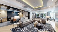 Located at 311 Karl Linn Drive in North Chesterfield, VA, this expansive single-floor amenity space offers a thoughtfully designed environment that blends social, recreational, and work-focused areas into one cohesive interior. The layout flows seamlessly between distinct zones, each defined by carefully curated furnishings and architectural details.

The main lounge area anchors the space with large-scale sectional sofas, a bold graphical area rug, and a striking large-format video display mounted against a dark textured accent wall. Herringbone-patterned hardwood flooring adds warmth and visual interest throughout the open areas. Overhead, tiered coffered ceilings with warm ambient LED cove lighting create a sense of depth and architectural elegance.

A dedicated gaming area features a full-size billiards table beneath a trio of modern pendant lights, with floor-to-ceiling windows drawing in abundant natural light and views of the surrounding outdoor spaces. Adjacent to this is a casual dining and lounge nook defined by exposed dark wood beam detailing, leather seating, and a wall-mounted display screen.

Glass-partitioned rooms along the perimeter provide private co-working or meeting spaces, maintaining visual connectivity with the broader area. A bar-height seating counter and coffee station add functional convenience. The overall ambiance is contemporary and upscale, balancing comfort with modern sophistication.