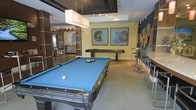 This single-floor game and social lounge is thoughtfully designed for recreation and entertainment. The centerpiece of the space is a full-sized billiards table with a vibrant blue felt surface, complemented by a shuffleboard table positioned along the back wall. A set of bar-height tables and sleek metal stools lines one side of the room, providing an ideal spot for spectators or casual conversation. Contemporary circular wall sculptures and a pair of bold, circular artwork prints in green and blue tones add visual interest to the yellow-toned accent walls. Overhead, a combination of a large rectangular pendant light and recessed track lighting ensures the space is well-lit throughout. Neutral carpeted flooring grounds the room, while warm wood-panel detailing on the columns and walls adds a refined, modern touch. A mirrored or glass-paneled partition visible in the background hints at additional adjacent spaces, enhancing the open and connected feel of the lounge. Overall, the space strikes a balance between a laid-back recreational atmosphere and a polished, contemporary aesthetic suitable for social gatherings.