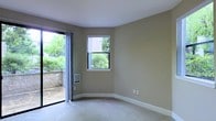 This single-floor space presents a clean, understated interior centered around a well-lit main room finished with neutral beige walls and wall-to-wall carpeting in a soft, light tone. White baseboard trim provides a crisp contrast to the warm wall color, contributing to a tidy, cohesive aesthetic throughout.

Natural light is a defining feature of the space. A double-hung window with a white frame offers a direct view of lush greenery outside, while a full-height sliding glass door with sheer panel curtains opens to an outdoor patio area. The outdoor space is bordered by mature shrubs and a low stone wall, creating a private, garden-like setting visible from inside.

A wall-mounted heating unit is positioned beneath the window, ensuring functional climate control without occupying floor space. Electrical outlets are conveniently placed along the walls. The room's open layout and dual sources of natural light give it an airy, versatile quality, making it adaptable to a range of uses. The overall ambiance is calm and neutral, well-suited for comfortable, everyday living.