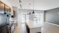 Located in Indianapolis's 46203 neighborhood, this two-story residence offers a thoughtfully designed interior with a clean, contemporary aesthetic. The main level features an open-concept kitchen and living area anchored by a large white waterfall island illuminated by three statement pendant lights. Rich dark wood cabinetry, black appliances, and track lighting complete the kitchen, while light hardwood flooring and an abundance of natural light from oversized windows and glass doors create a bright, airy atmosphere. The entryway makes a strong impression with a bold dark accent wall, built-in open shelving, and a sleek staircase with a modern cable-style railing. Upstairs, the bedrooms feature plush wall-to-wall carpet, large windows with tree-lined views, and ceiling fans for comfort. Multiple bathrooms are finished consistently with dark wood vanities, white countertops, light wood-look flooring, and clean white walls. One bathroom includes a shower-tub combination, while another offers a more open layout with a dedicated vanity area. Throughout both floors, the design balances modern industrial elements with warm, functional details, creating a cohesive and inviting living environment.