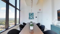This elegantly designed conference and meeting space occupies a high floor, immediately distinguished by its soaring ceilings and dramatic floor-to-ceiling windows that frame expansive city and treetop views. The crisp white walls and light neutral carpeting create a clean, bright backdrop that allows natural light to flood the interior throughout the day.

The primary conference area centers on a rich wood-toned rectangular table surrounded by contemporary black upholstered chairs with tapered wooden legs, offering comfortable seating for multiple attendees. A vibrant teal and white abstract painting adorns one wall, adding a curated artistic touch to the otherwise minimal palette. Pendant lighting descends from the lofty ceiling, providing focused illumination over the table.

An adjacent area features a wall-mounted flat-screen television positioned above a teal credenza styled with sculptural décor pieces, making it well-suited for presentations or collaborative work sessions. This zone flows openly into a secondary workspace area, visible through a wide architectural archway, where bar-height seating and additional work surfaces are arranged near more expansive windows.

Throughout, the space maintains a cohesive modern aesthetic, balancing functionality with refined design. The elevated vantage point, generous natural light, and seamless connection between areas make this a polished and productive environment.