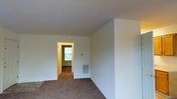 This single-floor residential space presents a straightforward and functional layout designed for comfortable everyday living. The interior is characterized by neutral, light-colored walls that create a bright and open atmosphere throughout. Wall-to-wall carpeting in a warm brown tone runs through the main living area, adding a sense of coziness underfoot. The space features standard ceiling heights that contribute to an airy feel. A wall-mounted HVAC vent provides efficient climate control directly within the living area. An interior doorway with classic wood trim connects to an adjacent room, which is highlighted by a window that allows natural light to filter in, offering views of the surrounding greenery outside. The clean, uncluttered design of the space provides a versatile canvas suitable for a variety of furniture arrangements and personal décor styles. Electrical outlets are positioned at regular intervals along the baseboards, ensuring practical utility throughout. Overall, the space combines simplicity and functionality, making it a solid foundation for a personalized and comfortable living environment.