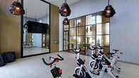 This contemporary fitness facility is thoughtfully designed to deliver both functionality and aesthetic appeal. The space is anchored by a dedicated spin cycling studio outfitted with multiple stationary bikes, making it ideal for high-energy indoor cycling sessions. Striking industrial-style pendant lights hang from the ceiling, adding a bold design element that complements the space's modern character.

One of the most notable architectural features is the expansive floor-to-ceiling steel-framed glass partition wall, which divides the cycling area from the adjacent fitness zones while maintaining an open, connected feel. This transparent boundary floods the interior with natural light and offers views of the surrounding outdoor greenery, creating an inviting atmosphere for workouts.

The flooring is finished in a clean, light-toned wood-look surface that enhances the brightness of the room. A large wall-mounted mirror on one side of the cycling studio amplifies the sense of space and allows exercisers to monitor their form. A flat-screen display is also mounted on the wall, supporting on-demand fitness content or instructional programming. The overall ambiance is sleek, energizing, and well-appointed, reflecting a high standard of design for a modern fitness environment.