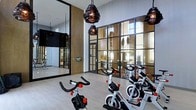 This contemporary fitness facility is thoughtfully designed to deliver both functionality and aesthetic appeal. The space is anchored by a dedicated spin cycling studio outfitted with multiple stationary bikes, making it ideal for high-energy indoor cycling sessions. Striking industrial-style pendant lights hang from the ceiling, adding a bold design element that complements the space's modern character.

One of the most notable architectural features is the expansive floor-to-ceiling steel-framed glass partition wall, which divides the cycling area from the adjacent fitness zones while maintaining an open, connected feel. This transparent boundary floods the interior with natural light and offers views of the surrounding outdoor greenery, creating an inviting atmosphere for workouts.

The flooring is finished in a clean, light-toned wood-look surface that enhances the brightness of the room. A large wall-mounted mirror on one side of the cycling studio amplifies the sense of space and allows exercisers to monitor their form. A flat-screen display is also mounted on the wall, supporting on-demand fitness content or instructional programming. The overall ambiance is sleek, energizing, and well-appointed, reflecting a high standard of design for a modern fitness environment.
