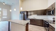 This well-designed single-floor unit showcases a contemporary aesthetic throughout, highlighted by dramatically high ceilings that lend an expansive, airy quality to the living spaces. Light-toned hardwood-style flooring flows seamlessly across the entire unit, creating a cohesive and clean foundation.

The open-concept kitchen is a standout feature, equipped with rich dark espresso cabinetry that extends nearly to the ceiling, granite countertops, and a full suite of stainless steel appliances including a refrigerator, range, microwave, and dishwasher. A white subway tile backsplash adds a modern touch, while a kitchen island with a granite surface and sleek metal legs provides additional prep space and a casual dining option. Pendant lighting hangs above the island, adding both function and style.

The living area is generously proportioned, benefiting from natural light and direct access to a private balcony through a sliding glass door, offering pleasant outdoor views of surrounding greenery. Stacked windows above the door further enhance the sense of height and light.

The bathroom features matching dark cabinetry with granite countertops, a well-lit mirror, and light wood-style flooring. An adjacent closet with wire shelving provides practical storage. Additional rooms include a bright secondary space with dual windows, well-suited for a bedroom or home office. Ceiling fans are present in the main living areas for added comfort.