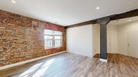 This thoughtfully designed single-floor residence seamlessly merges industrial character with modern finishes. Exposed red brick accent walls serve as a defining architectural feature throughout the space, appearing in the open living areas and bedrooms, adding warmth and historic texture to the interior. Dark wood ceiling beams and structural columns with decorative steel caps further reinforce the industrial aesthetic, while recessed lighting and crisp white walls maintain a clean, contemporary feel.

The main living area is bathed in natural light from large, multi-pane windows set directly into the brick, and durable light-toned hardwood-style flooring runs throughout the common spaces. The well-appointed kitchen features rich dark cabinetry extending to the ceiling for ample storage, light granite countertops, a stainless steel appliance suite including a range, microwave, dishwasher, and refrigerator, and continues the hardwood-style flooring for a cohesive look. In-unit laundry is also present.

The bedrooms offer plush carpeting for comfort, with one bedroom showcasing a charming arched brick window surround and ceiling fan. A walk-in closet and a transom window above the doorway add additional character. The bathroom is finished with classic white subway tile surrounding the soaking tub and shower combination, complemented by built-in shelving for convenient storage.