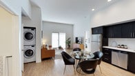 This single-floor residence is designed around an open-concept floor plan that seamlessly connects the kitchen, dining, and living areas into one cohesive, light-filled space. The interior is finished with light-toned hardwood-style flooring throughout, contributing to a clean and modern aesthetic complemented by crisp white walls and recessed lighting overhead.

The dining area is anchored by a round glass-top table surrounded by sleek black chairs with gold-toned legs, positioned adjacent to the kitchen. The kitchen features dark cabinetry, stainless steel refrigerator, and efficient use of vertical space. A stacked washer and dryer unit is conveniently integrated into a dedicated nook near the entryway, maximizing floor space.

The living area benefits from a large window that allows natural light to fill the room, creating a bright and airy atmosphere. A warm cognac-toned sofa, area rug, and decorative plants add texture and comfort to the space. A doorway visible in the background leads to a separate bedroom, providing a private retreat. The overall ambiance is contemporary, functional, and well-appointed.