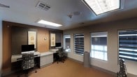 This single-floor workspace is designed with both functionality and comfort in mind. The room features two dedicated workstations, each equipped with computer monitors and ergonomic rolling chairs, making it well-suited for administrative or professional use. The built-in cabinetry beneath the desks provides practical storage, keeping the workspace organized and efficient.

The room's design balances warm and neutral tones, with a striking burnt orange accent wall contrasting against the cooler gray surroundings. Decorative framed artwork adorns the walls, adding a refined aesthetic to the professional environment. A pendant light fixture contributes to the layered lighting scheme alongside recessed ceiling lights.

Natural light is a notable feature, provided by multiple skylights in the ceiling and several windows fitted with adjustable horizontal blinds, allowing for precise light control. The windows also provide views to an adjacent interior space, suggesting this room may be part of a larger facility. The flooring is a warm-toned carpet, adding texture and a degree of acoustic comfort. A waste bin near the windows and a ceiling-mounted speaker and fire suppression system underscore the room's practical, professional character.