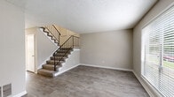 This two-story residence is finished in a consistent neutral palette, with warm beige walls, white trim, and textured ceilings throughout. The main level opens to a spacious living area with wood-look plank flooring and a carpeted staircase accented by a sleek metal railing leading to the upper floor. A second living or dining area on the main level features wall sconces, a chair rail detail, and sliding glass doors that open to a private outdoor balcony, allowing natural light to fill the room.

The kitchen is efficiently laid out with white cabinetry, a speckled quartz-style countertop, a gas cooktop, and white appliances including a refrigerator and dishwasher. A window above the sink brings in natural light and views of surrounding greenery.

Upstairs, the bedrooms are carpeted for comfort and receive natural light through blinded windows. One bedroom includes a ceiling fan and a walk-in closet. The upper level is served by two bathrooms: a full bath with a tiled tub-shower combination and a quartz vanity top, and a second bath with a single-sink vanity and wood-look tile flooring. The overall design is practical and move-in ready.