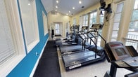 This fitness center is a dedicated, single-floor workout facility designed to support a comprehensive exercise routine. The space is thoughtfully laid out to maximize usability, featuring a row of treadmills along one side, complemented by elliptical machines and stationary bikes for a full range of cardio options. Toward the rear of the room, a multi-use strength training station provides opportunities for resistance and weight-based workouts.

The interior is bright and inviting, with large, multi-pane windows lining the walls and allowing abundant natural light to fill the space. Views of the surrounding greenery add a refreshing outdoor connection to the workout environment. Recessed lighting in the vaulted ceiling provides additional illumination, ensuring the space remains well-lit at any time of day.

The design features a neutral cream and bold teal color palette on the walls, giving the gym a clean, energetic aesthetic. Protective rubber flooring is installed beneath the equipment, prioritizing safety and durability. A mounted television is also present, adding an entertainment option for users during workouts. The overall ambiance is functional, clean, and welcoming, making it a practical and pleasant space for daily fitness activities.